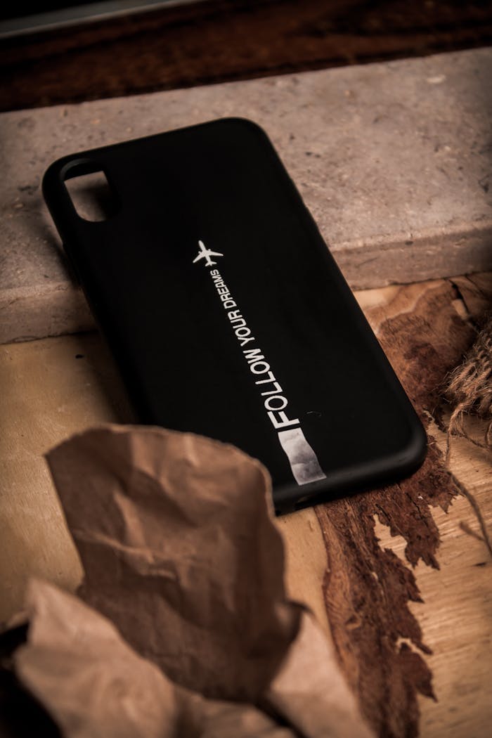 A close-up of a black phone case with 'Follow Your Dreams' text lying on a rustic surface.